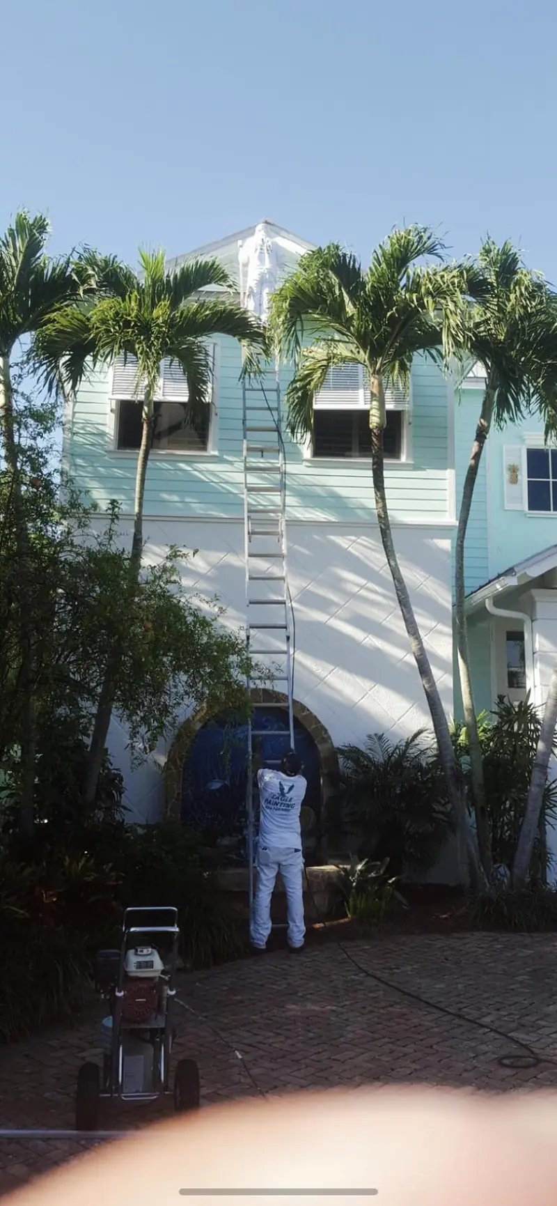Painter on an extension ladder repainting a two-story Broward house with palm trees